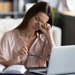 Zmęczona kobieta pociera oko, szukając sposobu, jak nawilżyć oczy bez kropli. Obok laptop i otwarty notatnik.
