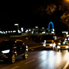 Nocna jazda przez miasto, gdzie rozmazane światła samochodów i latarni tworzą magiczną poświatę. W oddali widać niebieskie koło diabelskiego młyna.