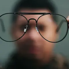 Osoba trzyma przed twarzą okulary, przez które świat wydaje się dziwnie zniekształcony. Nowe okulary, dziwne widzenie.