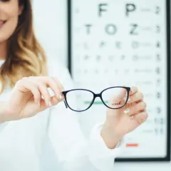Okulary z astygmatyzmem – czy trzeba nosić cały czas? Optometrystka prezentuje okulary na tle tablicy Snellena.