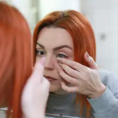 Kobieta z rudymi włosami zakłada soczewkę kontaktową, zastanawiając się, dlaczego soczewki zachodzą mgłą.