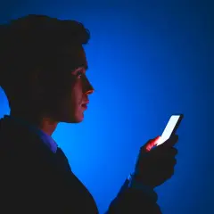 Mężczyzna w garniturze patrzy na świecący ekran telefonu. Jasne światło telefonu odbija się w jego oku, jakby chroniło je przed ciemnością.