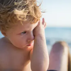 Chłopiec z piaskiem w oczach, zasłaniający je ręką. W tle morze. Uczucie piasek w oczach domowe sposoby.