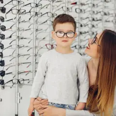 Chłopiec w okularach, które wydają się za małe, stoi obok mamy w salonie optycznym.