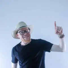 Uśmiechnięty albinos człowiek w kapeluszu i okularach wskazuje palcem do góry.