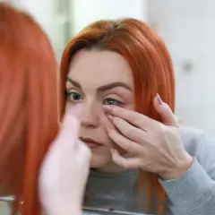 Ruda kobieta w lustrze zakłada soczewkę. Czy zakładanie soczewek boli? Nie, jeśli robi się to delikatnie.