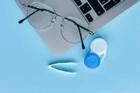 Okulary, pojemnik na soczewki i pęseta na laptopie. Czy można nosić soczewki i okulary jednocześnie? Tak, jeśli potrzebujesz korekcji wzroku.