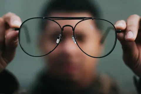 Osoba trzyma przed twarzą okulary, przez które świat wydaje się dziwnie zniekształcony. Nowe okulary, dziwne widzenie.