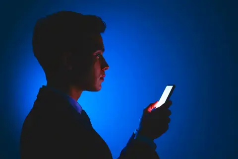 Mężczyzna w garniturze patrzy na świecący ekran telefonu. Jasne światło telefonu odbija się w jego oku, jakby chroniło je przed ciemnością.