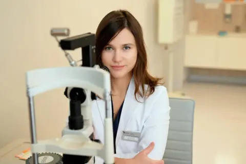 Okulistka w białym kitlu przy aparaturze. Czeka na pacjenta, któremu poda krople u okulisty.