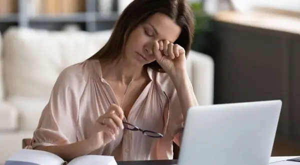 Zmęczona kobieta pociera oko, szukając sposobu, jak nawilżyć oczy bez kropli. Obok laptop i otwarty notatnik.
