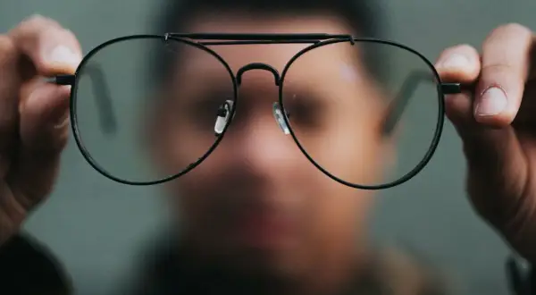 Osoba trzyma przed twarzą okulary, przez które świat wydaje się dziwnie zniekształcony. Nowe okulary, dziwne widzenie.