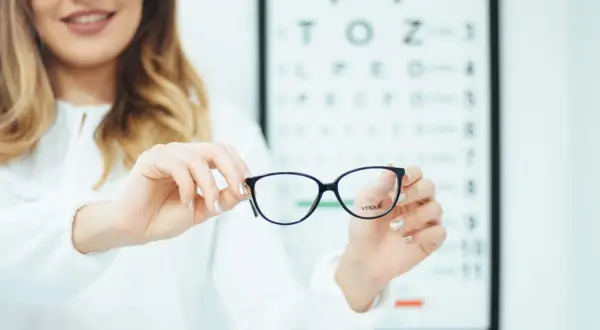 Okulary z astygmatyzmem – czy trzeba nosić cały czas? Optometrystka prezentuje okulary na tle tablicy Snellena.