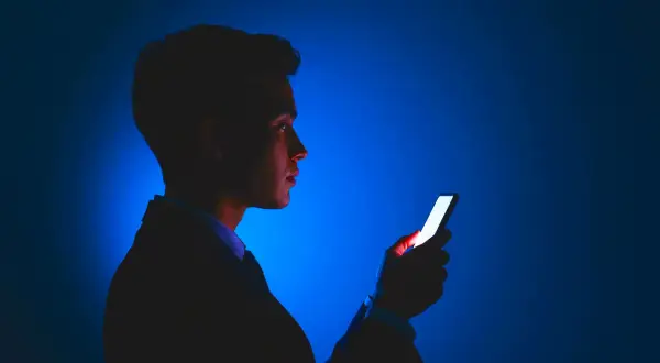 Mężczyzna w garniturze patrzy na świecący ekran telefonu. Jasne światło telefonu odbija się w jego oku, jakby chroniło je przed ciemnością.
