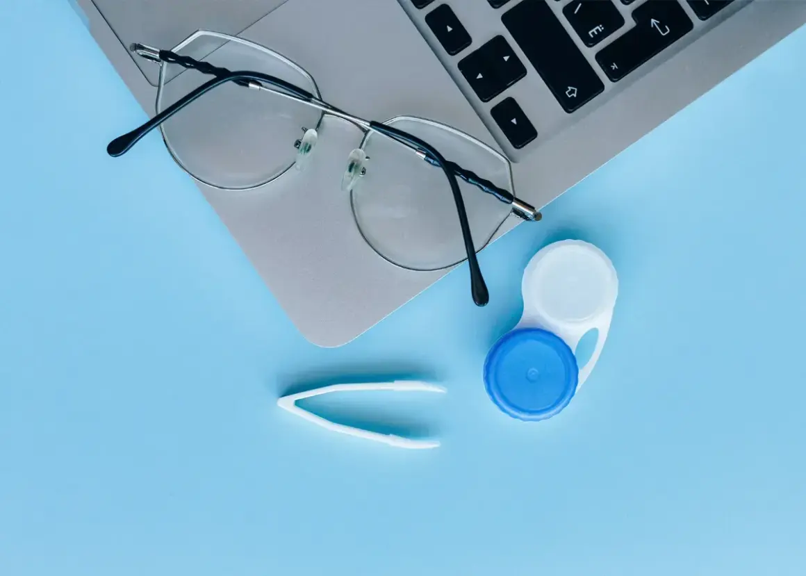 Okulary, pojemnik na soczewki i pęseta na laptopie. Czy można nosić soczewki i okulary jednocześnie? Tak, jeśli potrzebujesz korekcji wzroku.