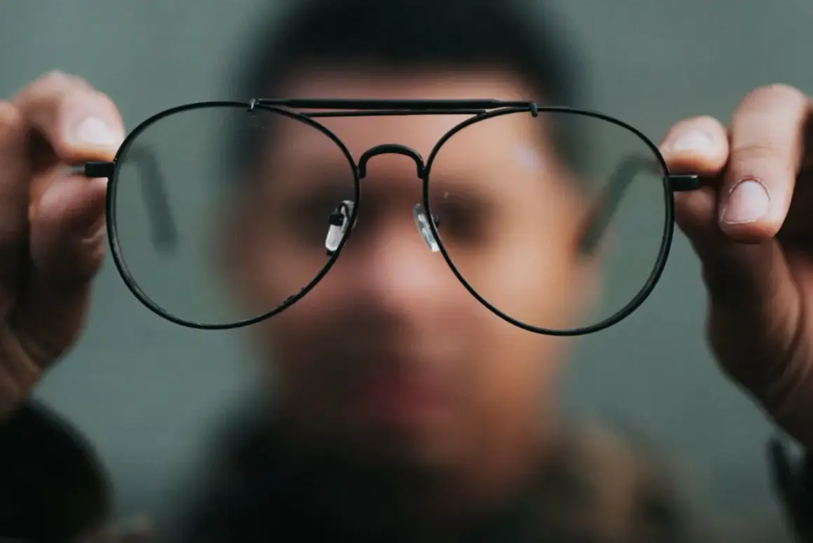 Osoba trzyma przed twarzą okulary, przez które świat wydaje się dziwnie zniekształcony. Nowe okulary, dziwne widzenie.