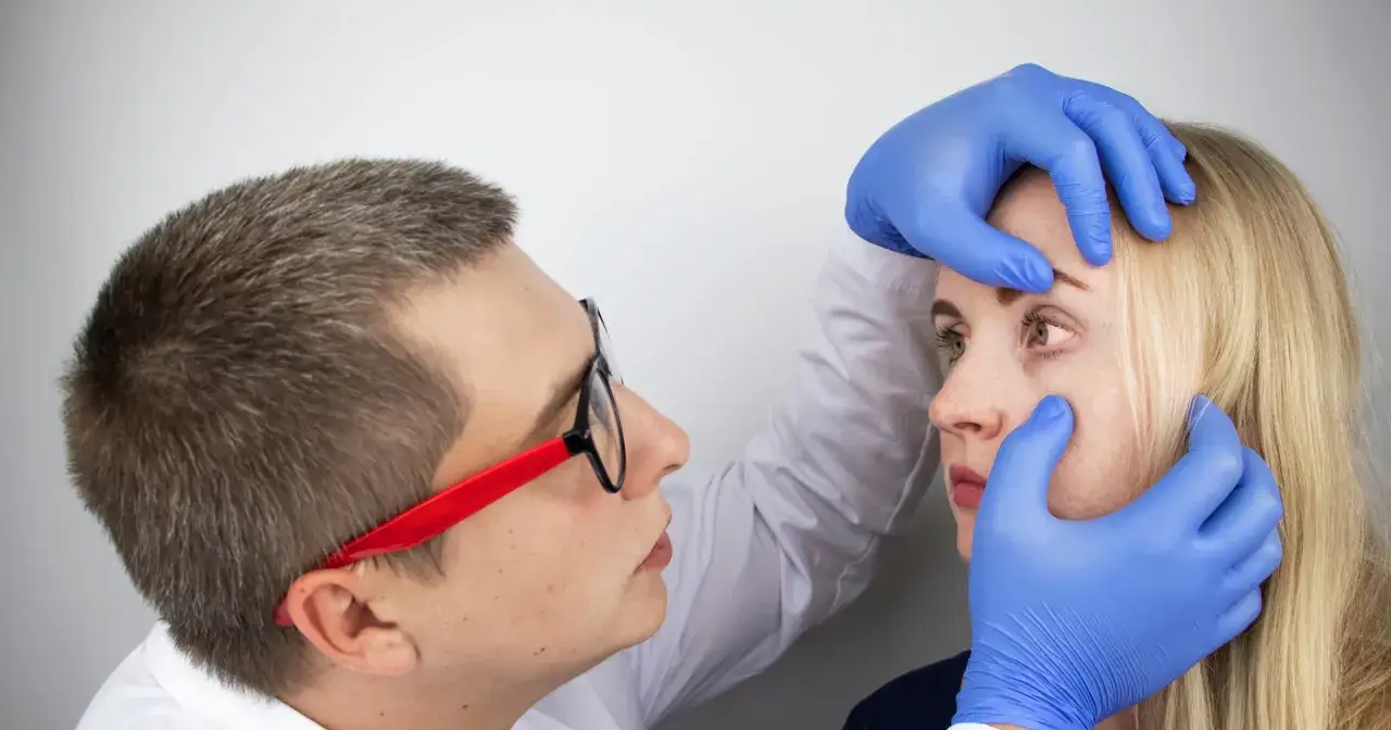 Lekarz w niebieskich rękawiczkach bada oko pacjentki. Może to być etap diagnozy zapalenia rogówki, zanim rozpocznie się leczenie domowe.