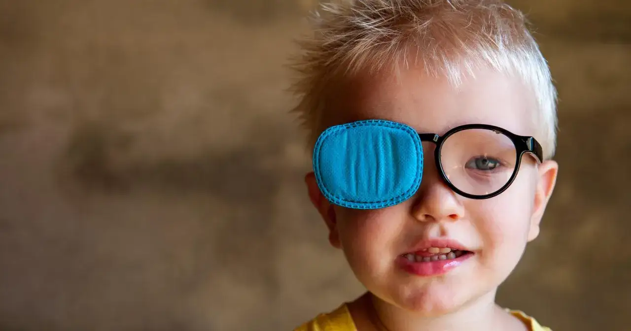 Mały chłopiec z niebieską łatką na oku, kt&oacute;ra ukrywa zez. Nosi okulary, a jego uśmiech jest pełen radości.