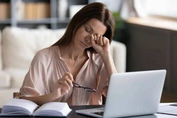 Zmęczona kobieta pociera oko, szukając sposobu, jak nawilżyć oczy bez kropli. Obok laptop i otwarty notatnik.
