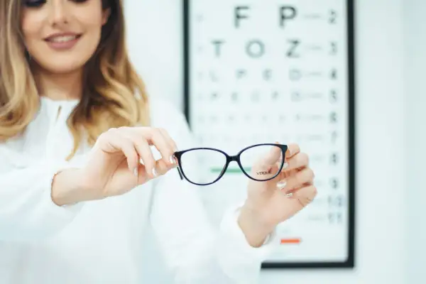 Okulary z astygmatyzmem – czy trzeba nosić cały czas? Optometrystka prezentuje okulary na tle tablicy Snellena.