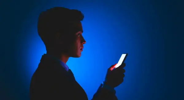 Mężczyzna w garniturze patrzy na świecący ekran telefonu. Jasne światło telefonu odbija się w jego oku, jakby chroniło je przed ciemnością.